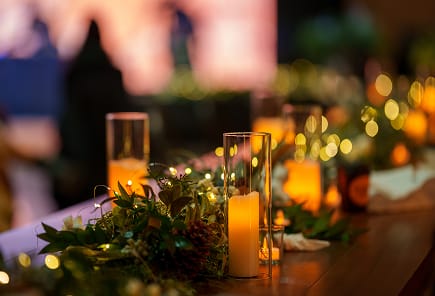 A close-up of a warmly lit table decorated for an event, featuring glass candle holders with glowing candles, surrounded by greenery and small twinkling lights.