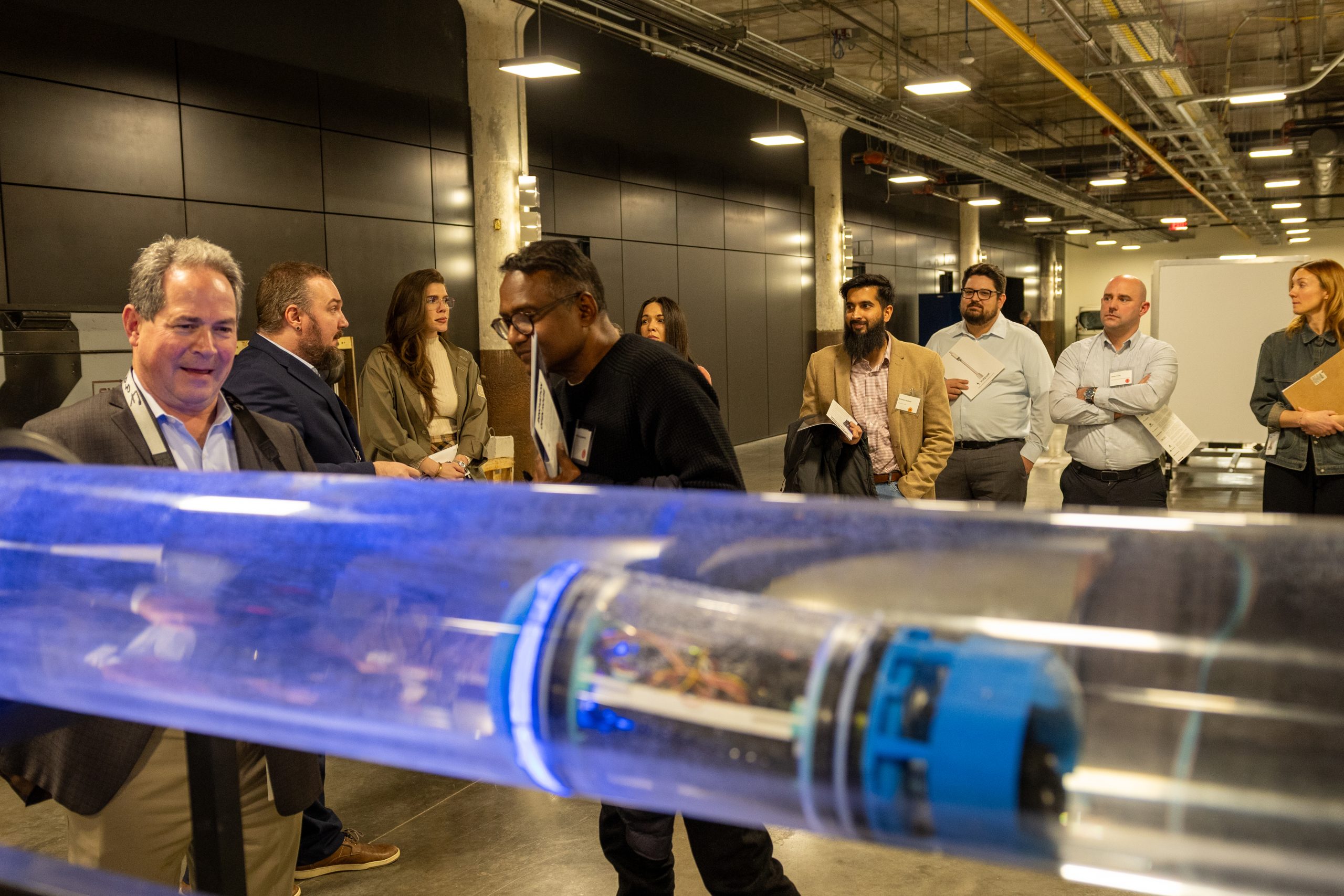 A group of professionals stands in an industrial space, attentively watching a demonstration of a Motmot autonomous inspection robot. The blue and white cylindrical robot is visible inside a large, clear horizontal tube filled with water in the foreground, simulating a pipeline inspection, while observers look on with interest.
