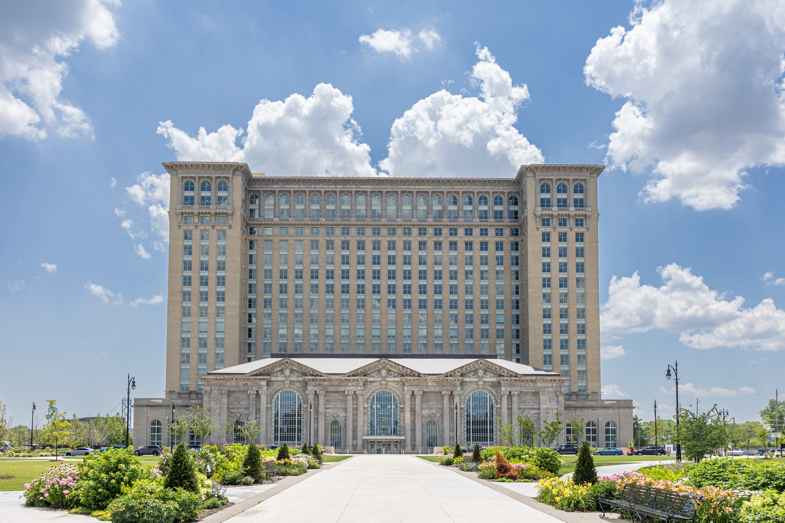 A straight-on, eye-level view of the Michigan Central Station under a bright blue sky with fluffy white clouds. The massive historic building features a tall, rectangular office tower with rows of windows rising from a grand, ornate base with three large arched windows and classical columns. A wide paved walkway leads symmetrically to the main entrance, flanked on both sides by landscaped gardens filled with blooming flowers, green shrubs, and young trees.