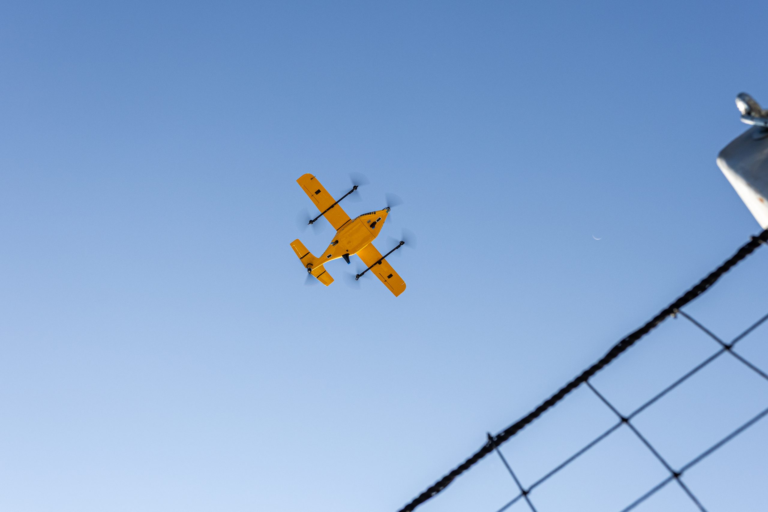 A bright yellow fixed-wing drone with four spinning rotors flies overhead against a clear, deep blue sky. The underside of the aircraft is visible as it moves through the air, framed by a blurred chain-link fence in the bottom right corner.