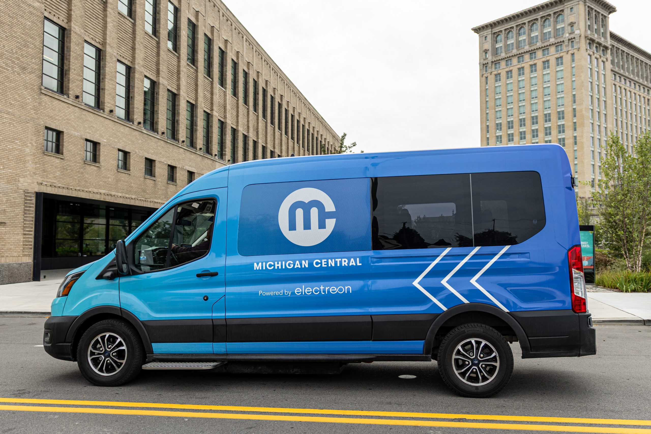 A side profile view of a bright blue Electreon shuttle van on a city street. The vehicle features a Michigan Central logo and the text "Powered by Electreon," along with white chevron graphics on the rear panel. The historic Michigan Central Station tower and the Book Depository building where Newlab Detroit is located are visible in the background.