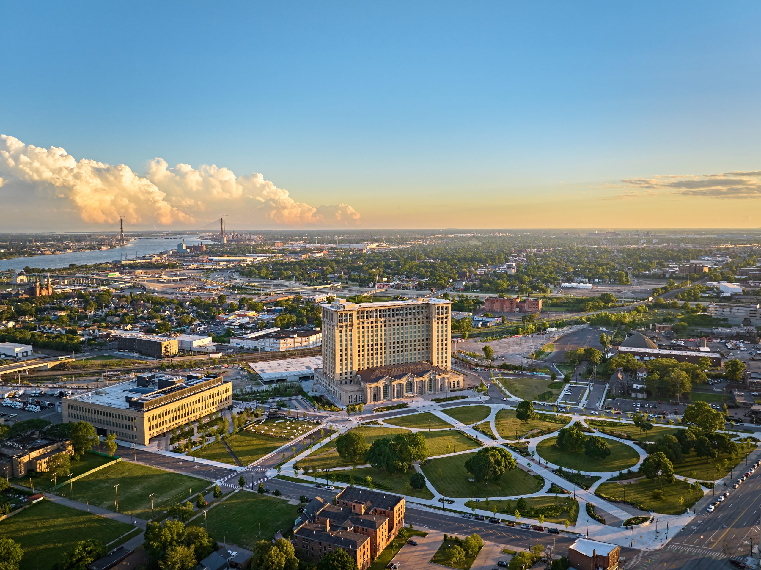 An aerial view of the Michigan Central Station, a large, ornate, light-colored building, surrounded by newly landscaped green parks with winding paths and trees. In the foreground, other buildings and roads are visible. In the background, a cityscape extends towards the horizon, with the Detroit River and the partially constructed Gordie Howe International Bridge visible under a bright blue sky with large, warm-toned clouds.