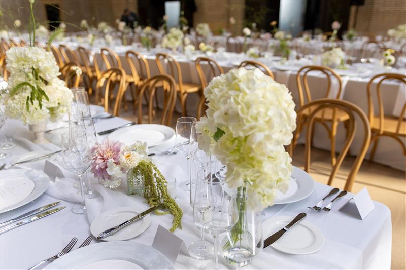 A long dining table set with white tablecloths, white plates, clear glassware, folded place cards, and tall glass vases holding white floral arrangements, with rows of curved wooden chairs extending into the background.