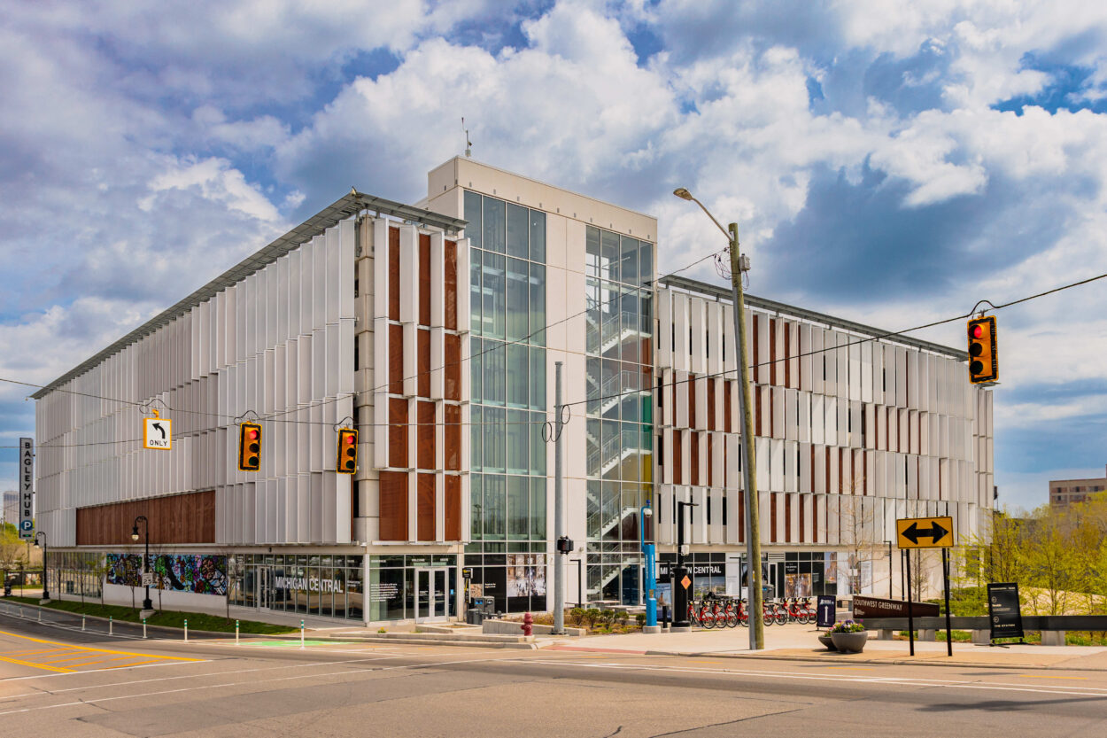 Exterior view of a modern multi-story building with alternating panels of wood cladding and floor-to-ceiling glass windows under a partly cloudy sky, with a street and traffic light visible in the foreground.