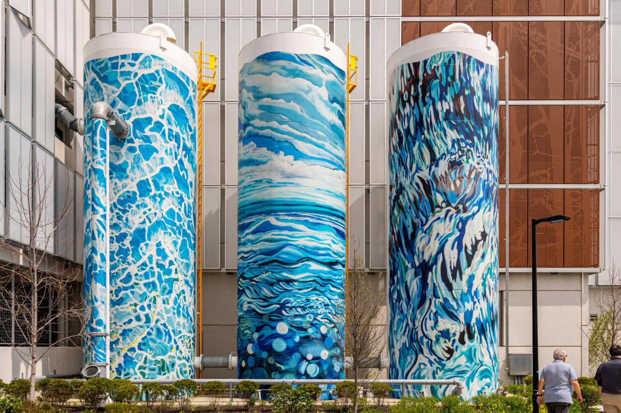 Three large cylindrical silos painted with vibrant blue-and-white fluid art depicting swirling water or wave patterns, displayed outdoors.