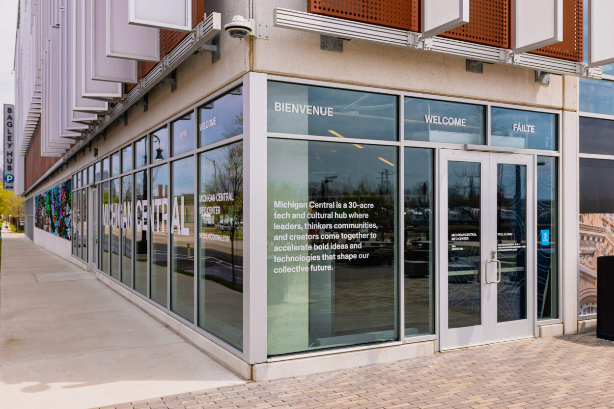 Exterior entrance of a modern commercial or civic building with floor-to-ceiling glass storefront windows, displaying green text signage, with a covered walkway and street visible outside.