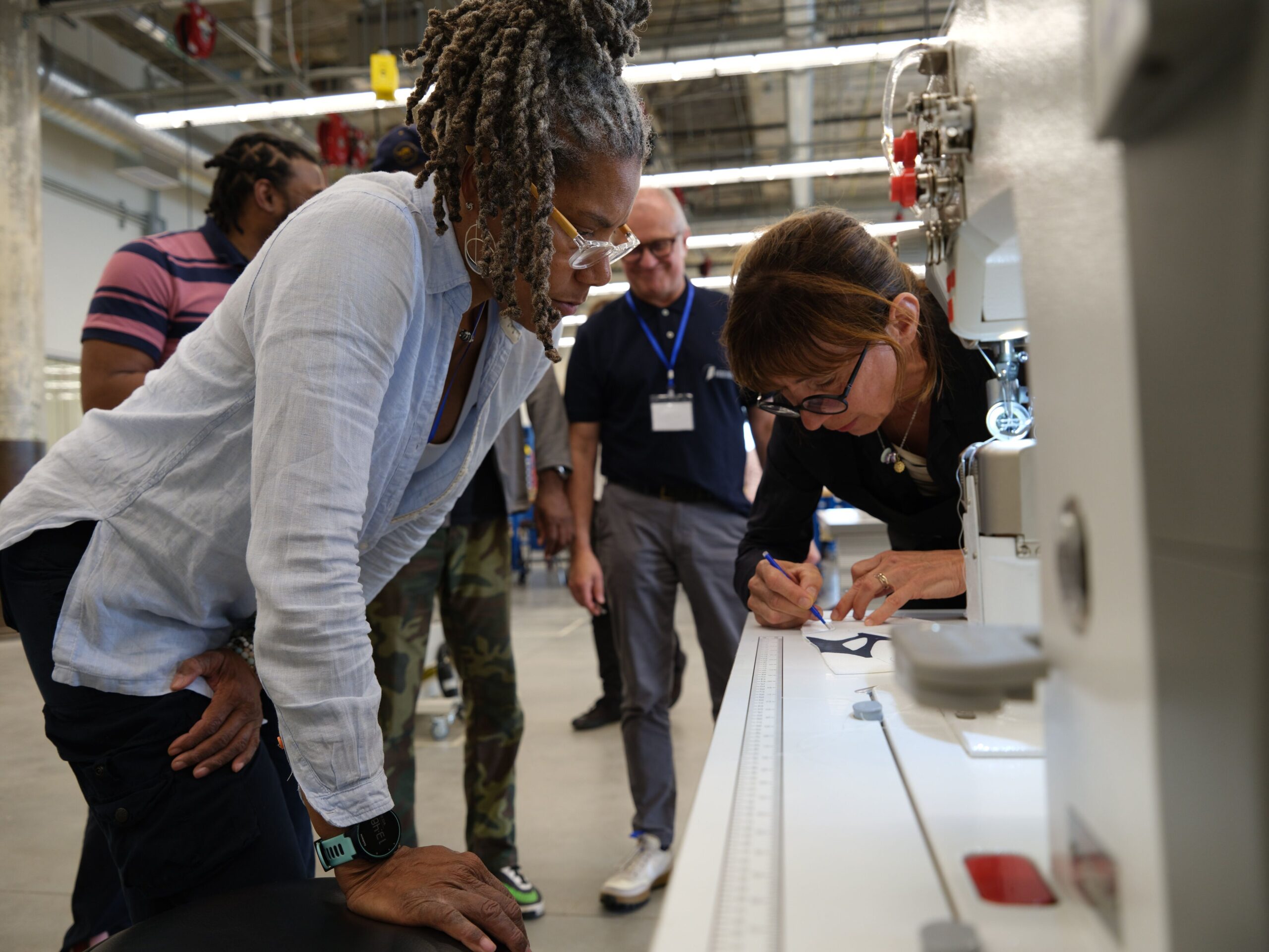 Two women examine a fabric pattern at an industrial machine, while colleagues look on.