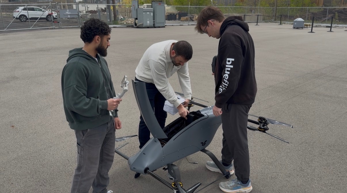 Three people assemble or inspect a large gray multirotor drone/eVTOL aircraft in an outdoor paved area. One person wears a "Blueflight" branded hoodie.