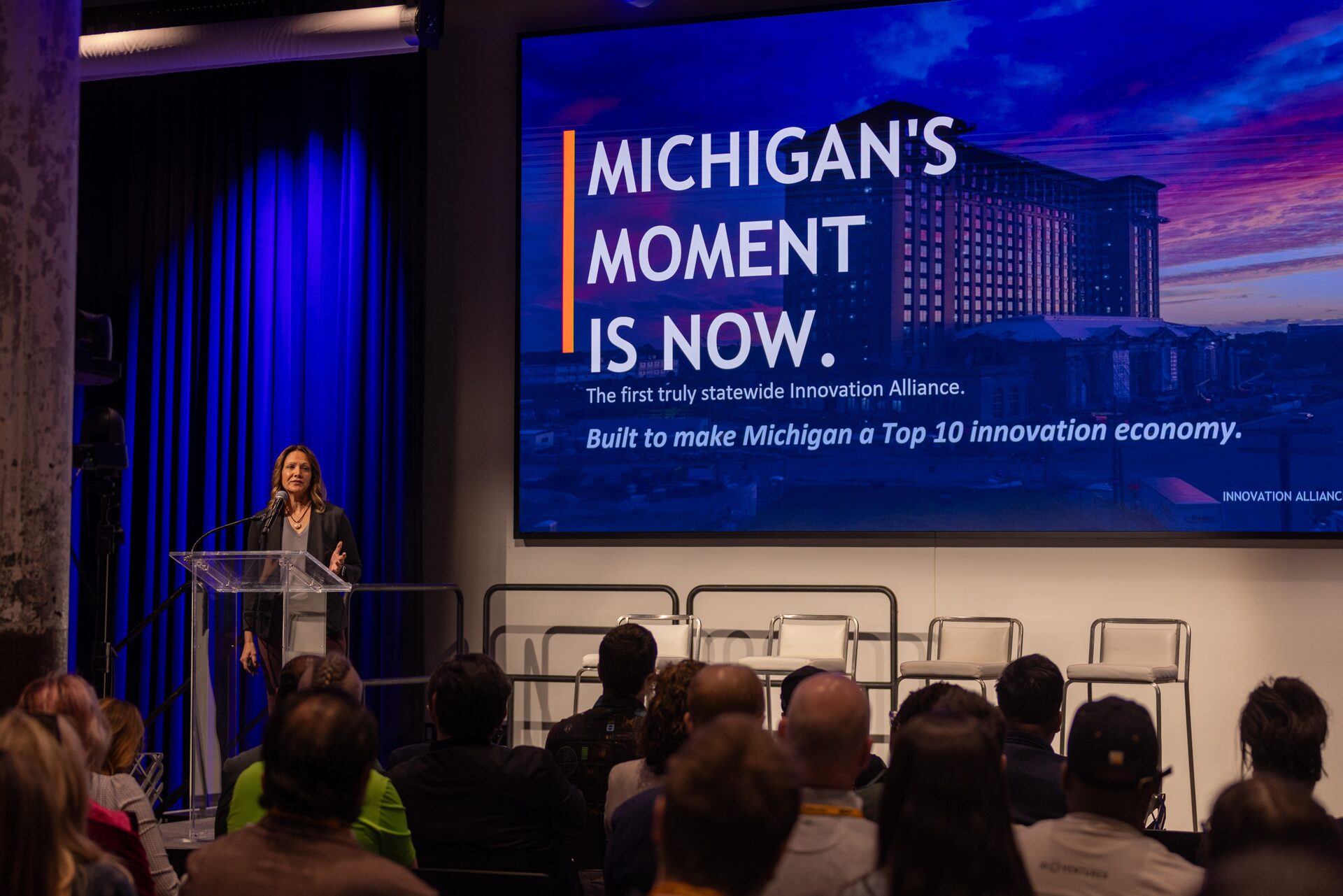 A speaker presents at a podium before an audience at a Michigan Innovation Alliance event. A large screen behind her displays the message "Michigan's Moment Is Now — The first truly statewide Innovation Alliance, built to make Michigan a Top 10 innovation economy."