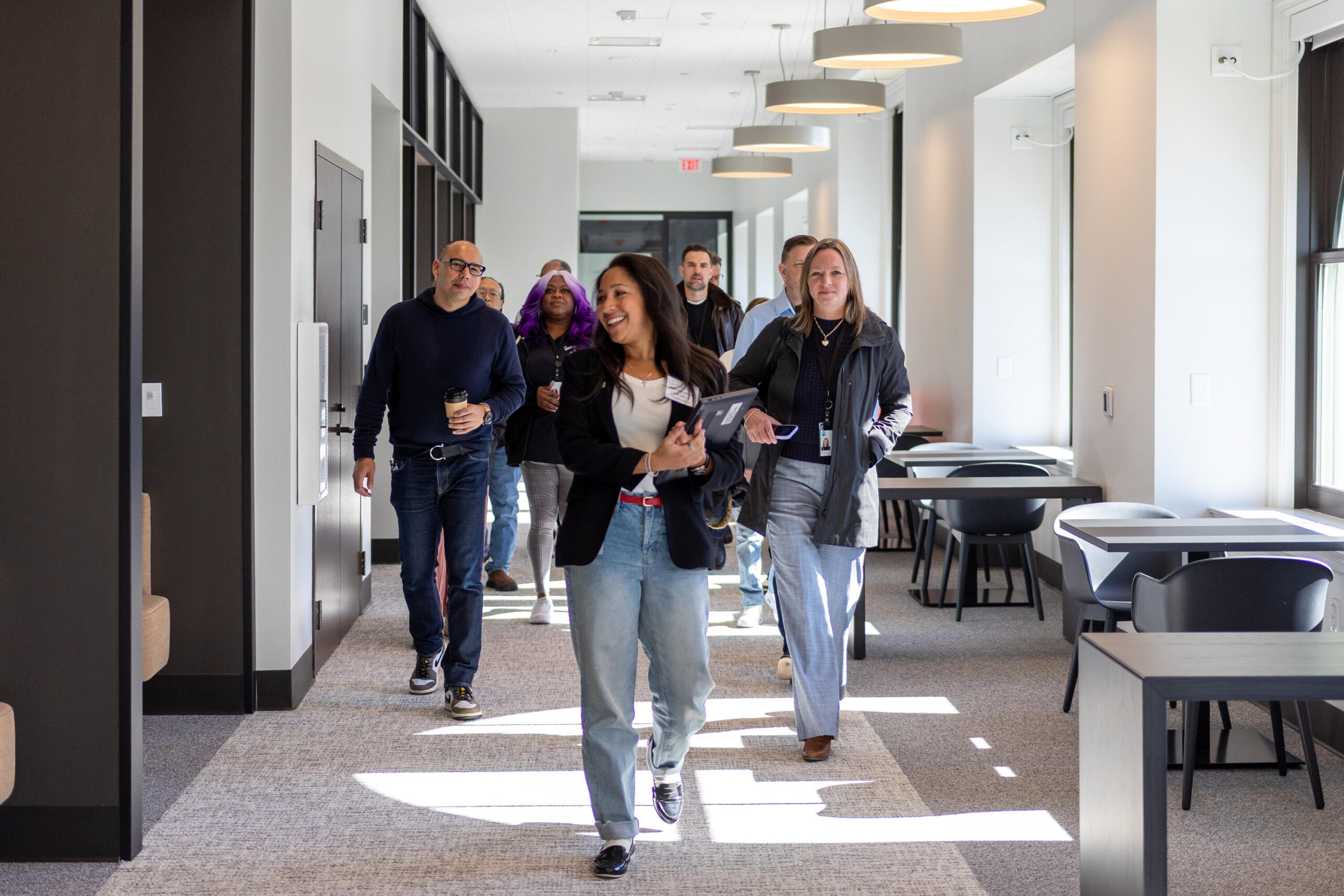A group of people walks through a bright, modern office corridor during a tour or opening day event. A woman in the foreground smiles warmly while holding a tablet, leading the group past open workspace seating bathed in natural light from large windows. Pendant lights line the ceiling above.