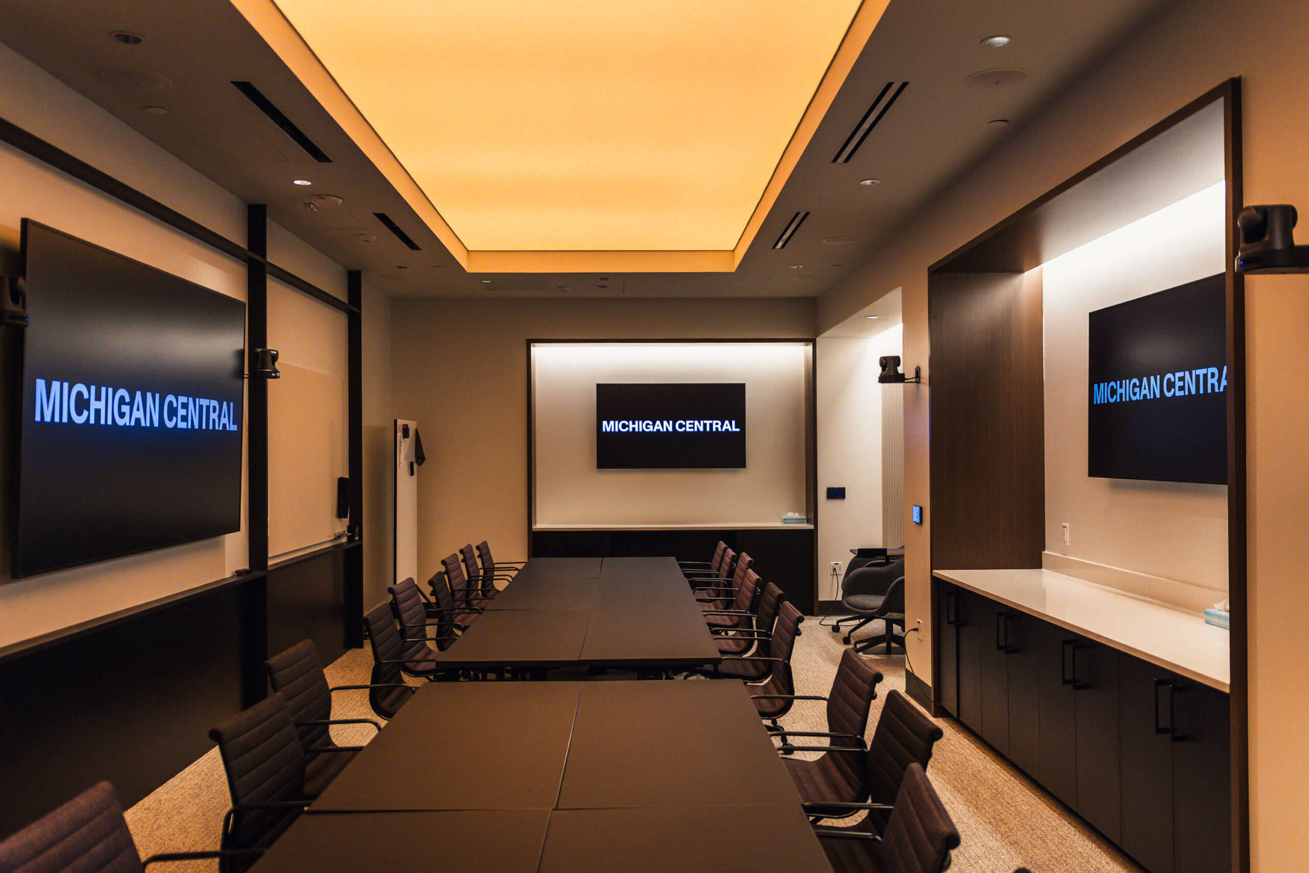 A professional boardroom at Michigan Central featuring a long dark conference table surrounded by upholstered chairs. Three large screens display the "Michigan Central" wordmark in blue. The room has warm ambient lighting from a backlit ceiling panel, dark wood accents, and video conferencing cameras mounted on the walls.
