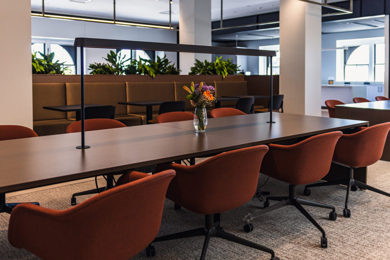 A modern collaborative workspace featuring a long dark wood conference table centered under a sleek linear pendant light, surrounded by terracotta-upholstered chairs on casters. A small vase of colorful flowers sits at the center of the table. In the background, booth-style seating is lined with lush greenery along the windows, creating a warm and inviting atmosphere.