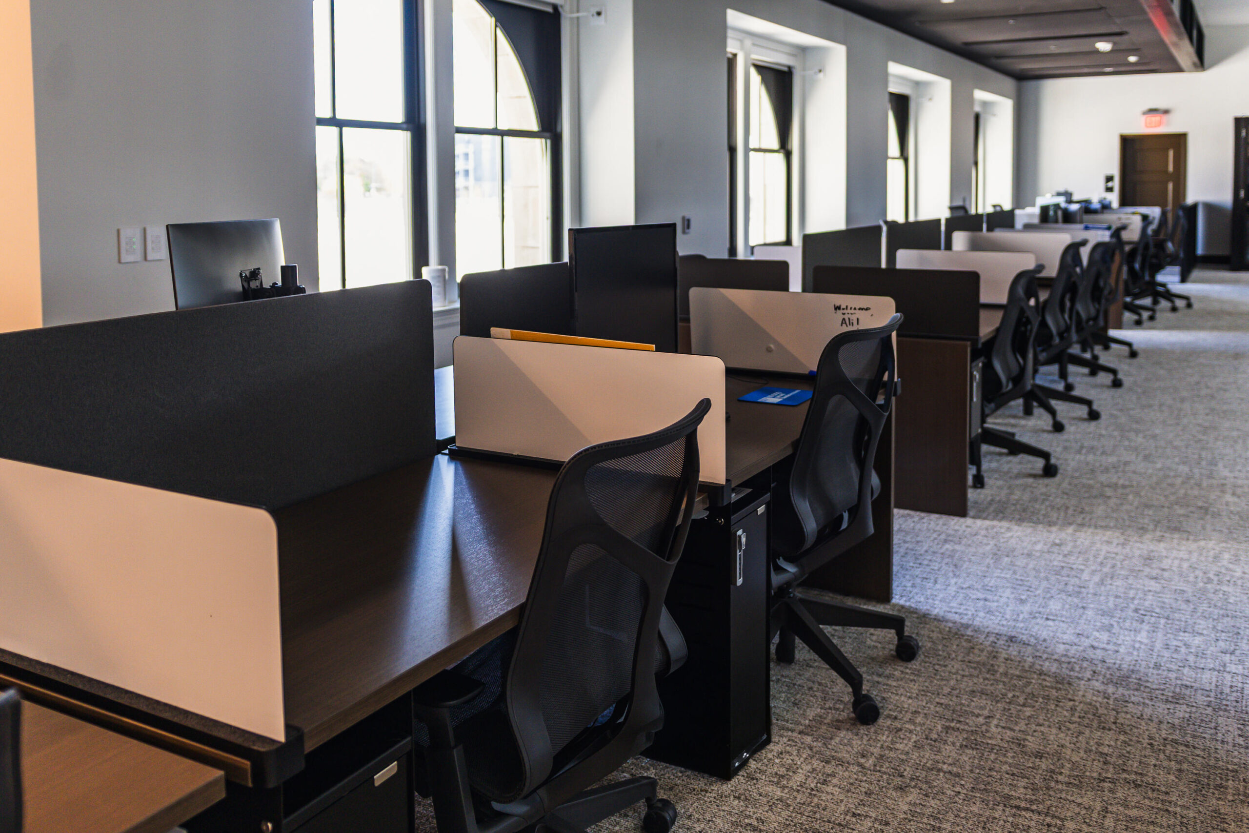 Rows of open-plan workstations with dark wood desks, privacy dividers, and ergonomic mesh chairs stretch down a bright office floor. Monitors sit atop each station, and large arched windows along the wall fill the space with natural light.